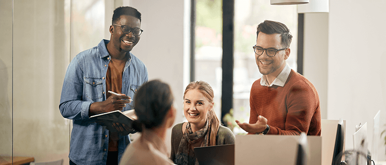 Group of coworkers smiling and collaborating in a modern office, discussing ideas around desks with laptops and notebooks.