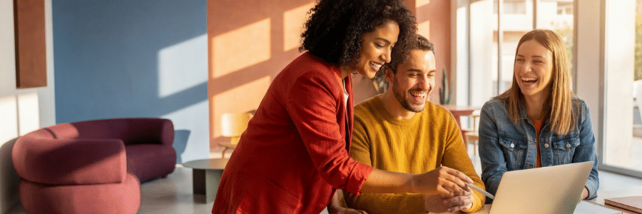 Photograph of three people collaborating around a laptop in a brightly lit modern office with large windows and colorful furniture. One person in a red jacket is pointing at the laptop screen while the other two, wearing yellow and denim jackets, attentively observe.